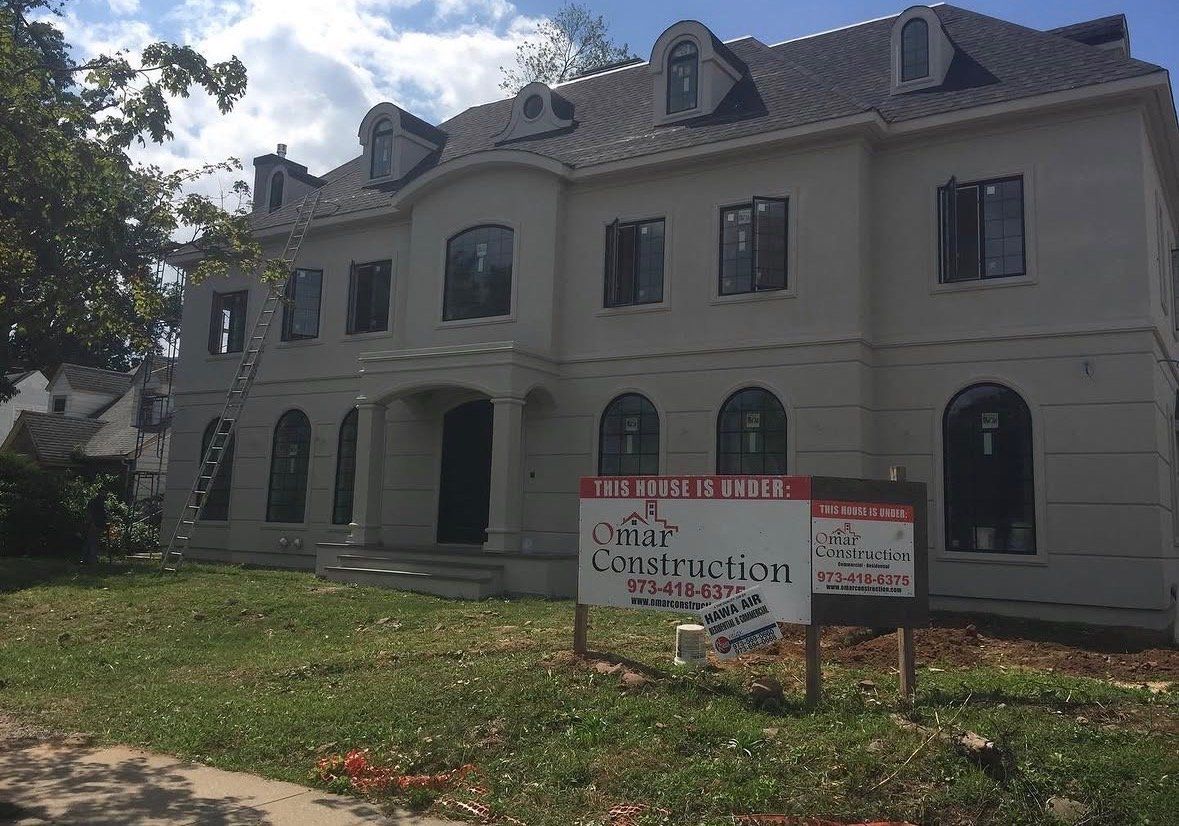 New two-story stucco house with arched windows and dormers under construction, with an Omar Construction project sign on the lawn